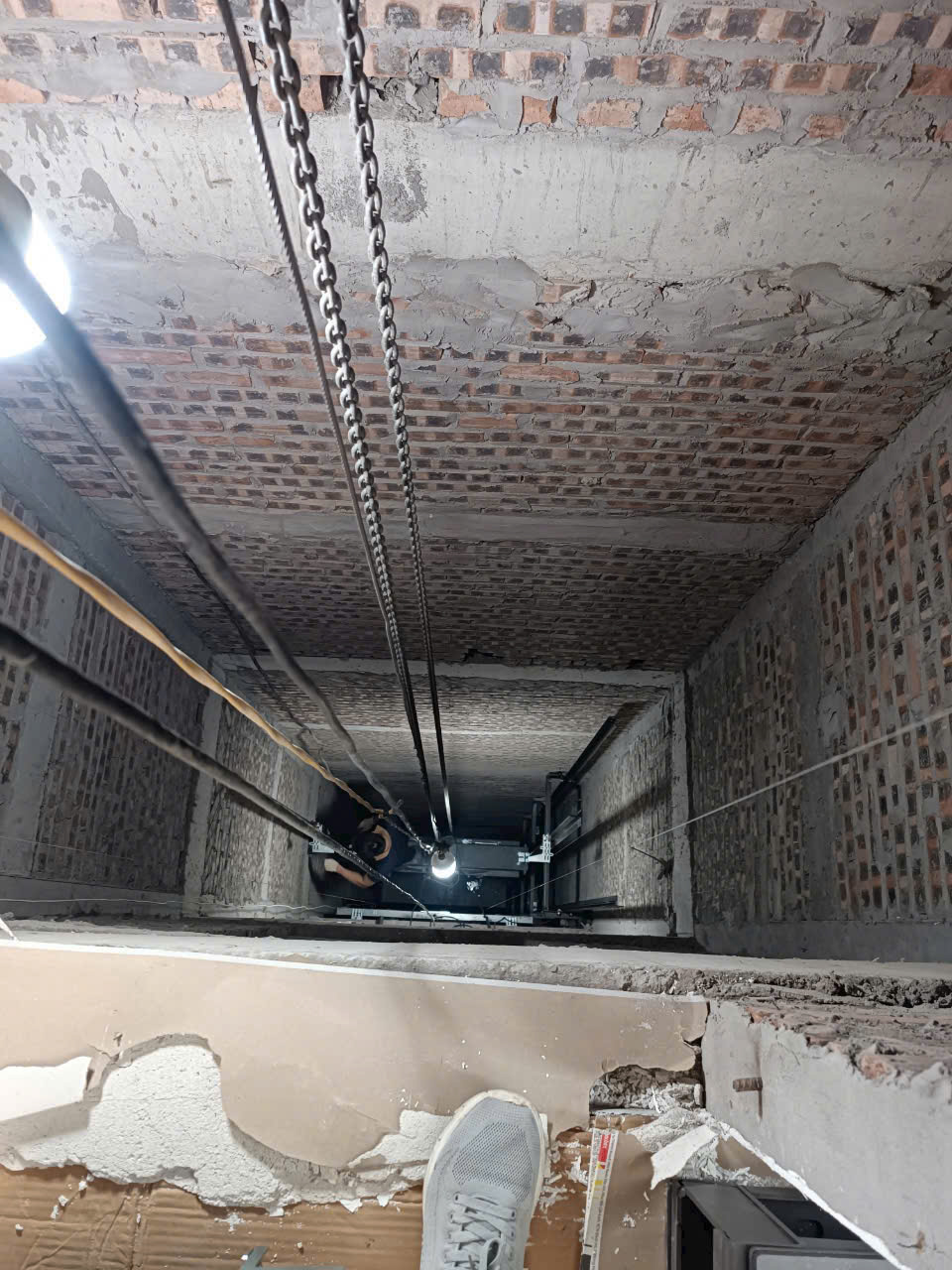 Hố thang máy đang thi công với hệ thống cáp xích và tường gạch thô nhìn từ trên xuống